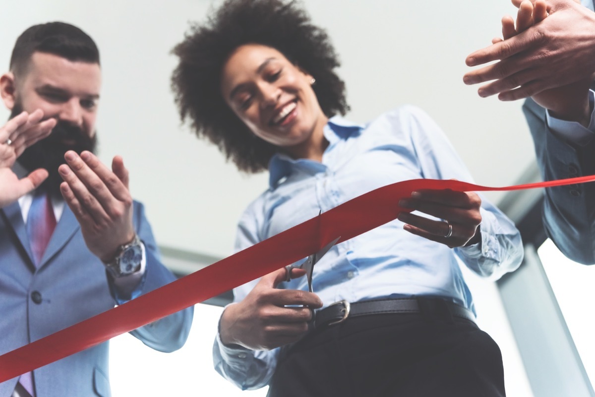 Young beautiful ambitious cheerful mulatto businesswoman cutting red ribbon on a ceremonial grand opening of startup company while her associates clapping hands and celebrating