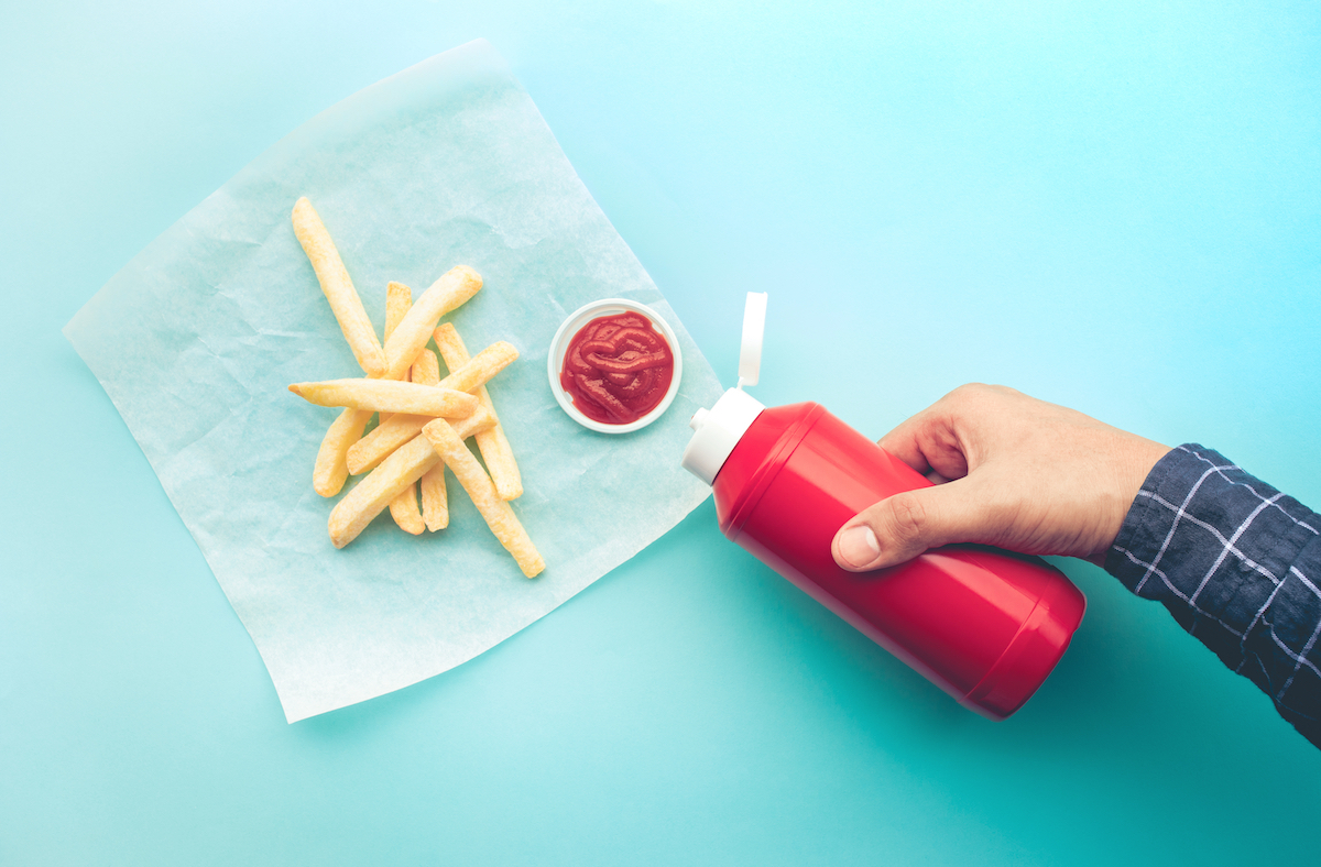 Hand pouring ketchup in cup next to fries