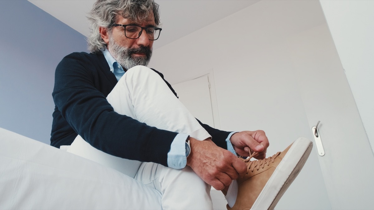 Mature man putting on his casual comfort shoes in front of bed, tying his shoelace