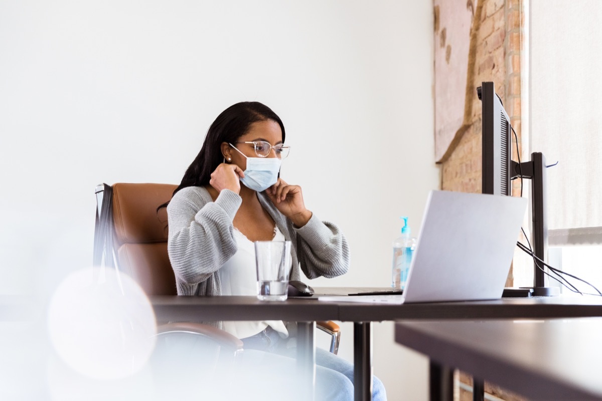 A mid adult woman puts on a protective mask as she starts working in her office. She is looking at something on a laptop. She is working in an office amid the COVID-19 pandemic.