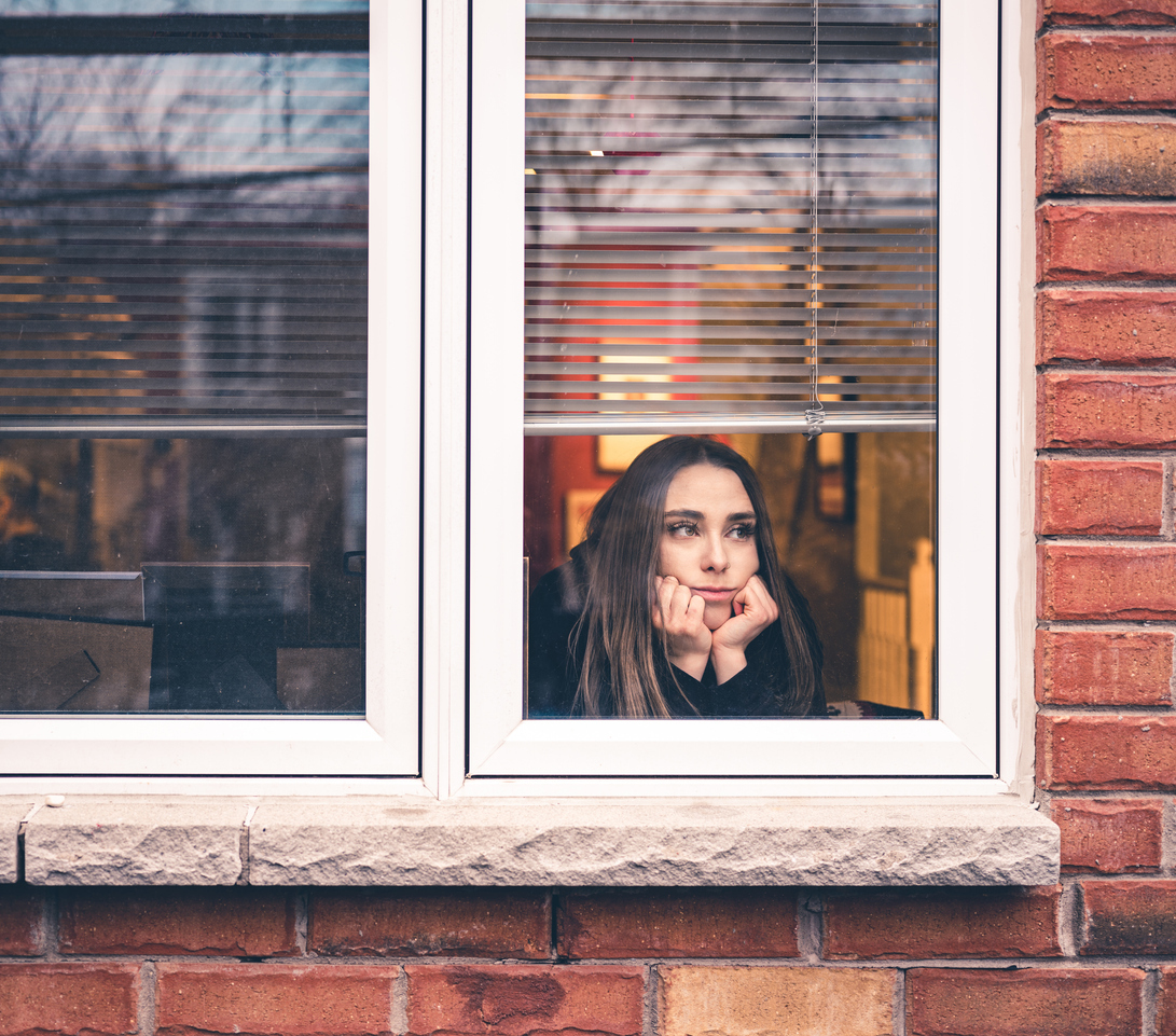 A young woman looks out her window with a distressed look on her face due to coronavirus lockdowns