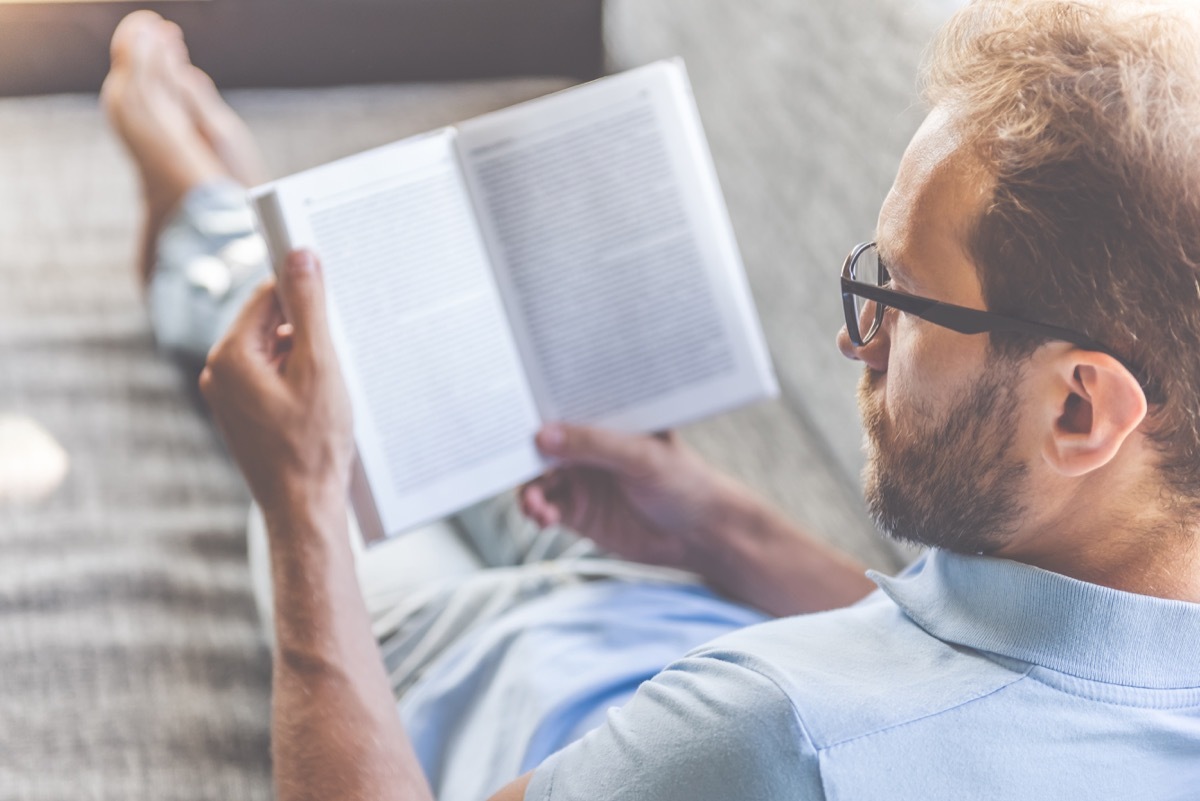 Man in casual clothes and eyeglasses reading a book while lying on couch at home