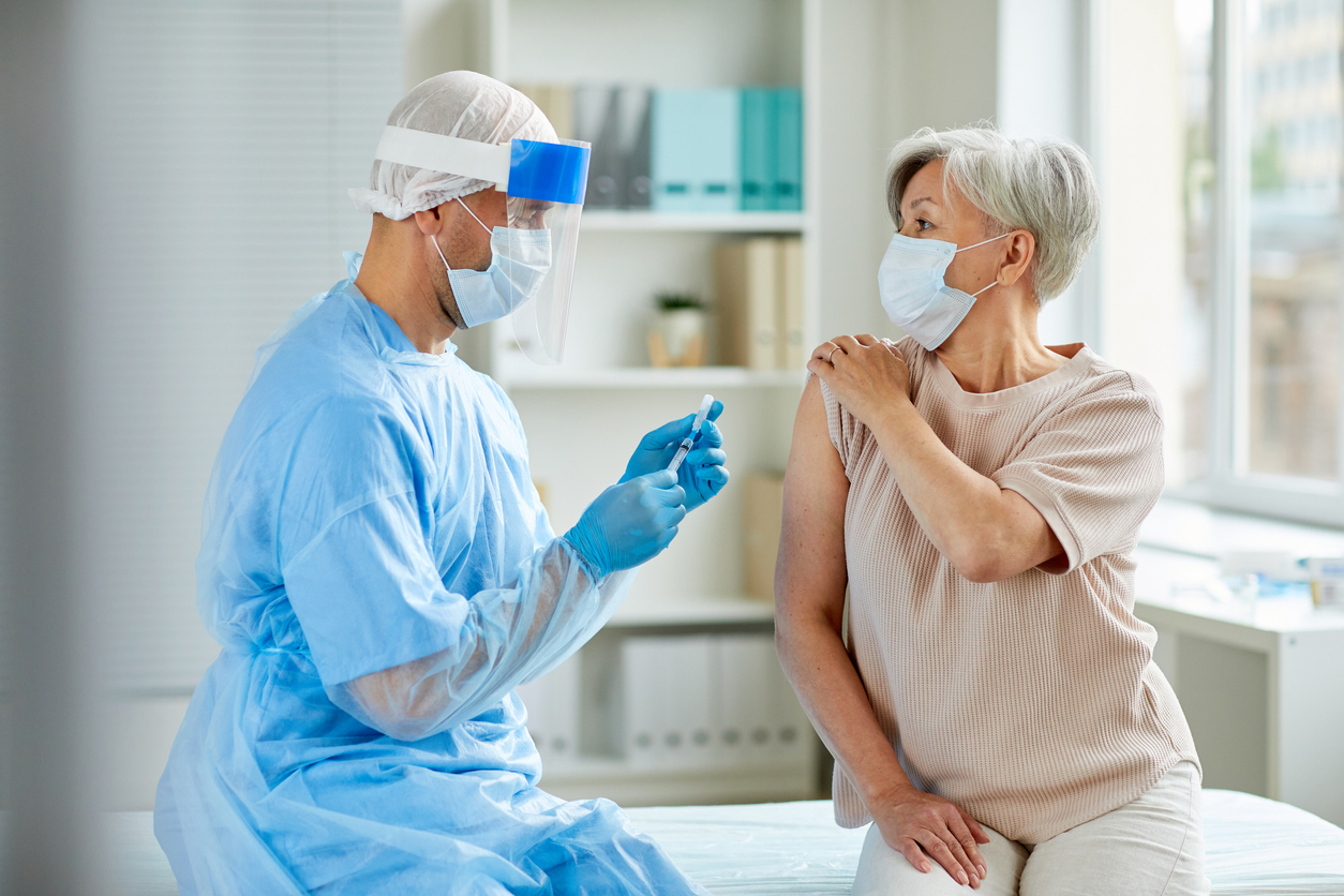 An older woman pulling up her sleeve to get a COVID vaccine from a male health care worker wearing PPE.