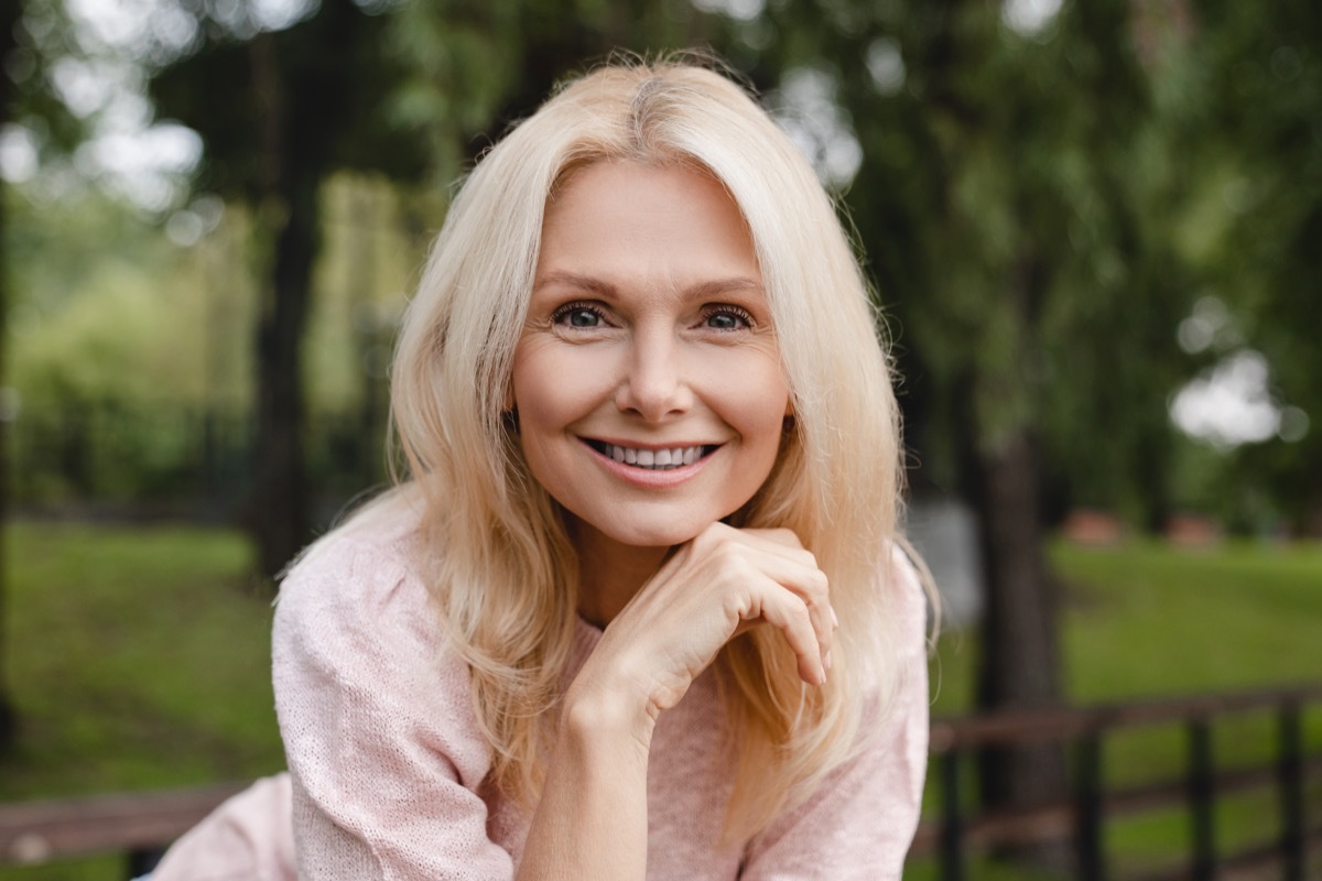 Cheerful mature blonde woman wife with toothy smile relaxing resting walking in city park forest alone looking at camera outdoors