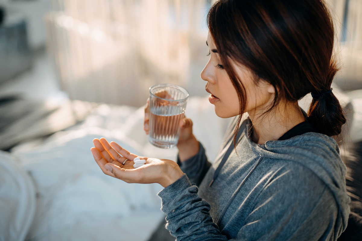 Young woman sitting on bed and feeling sick, taking medication in hand with a glass of water