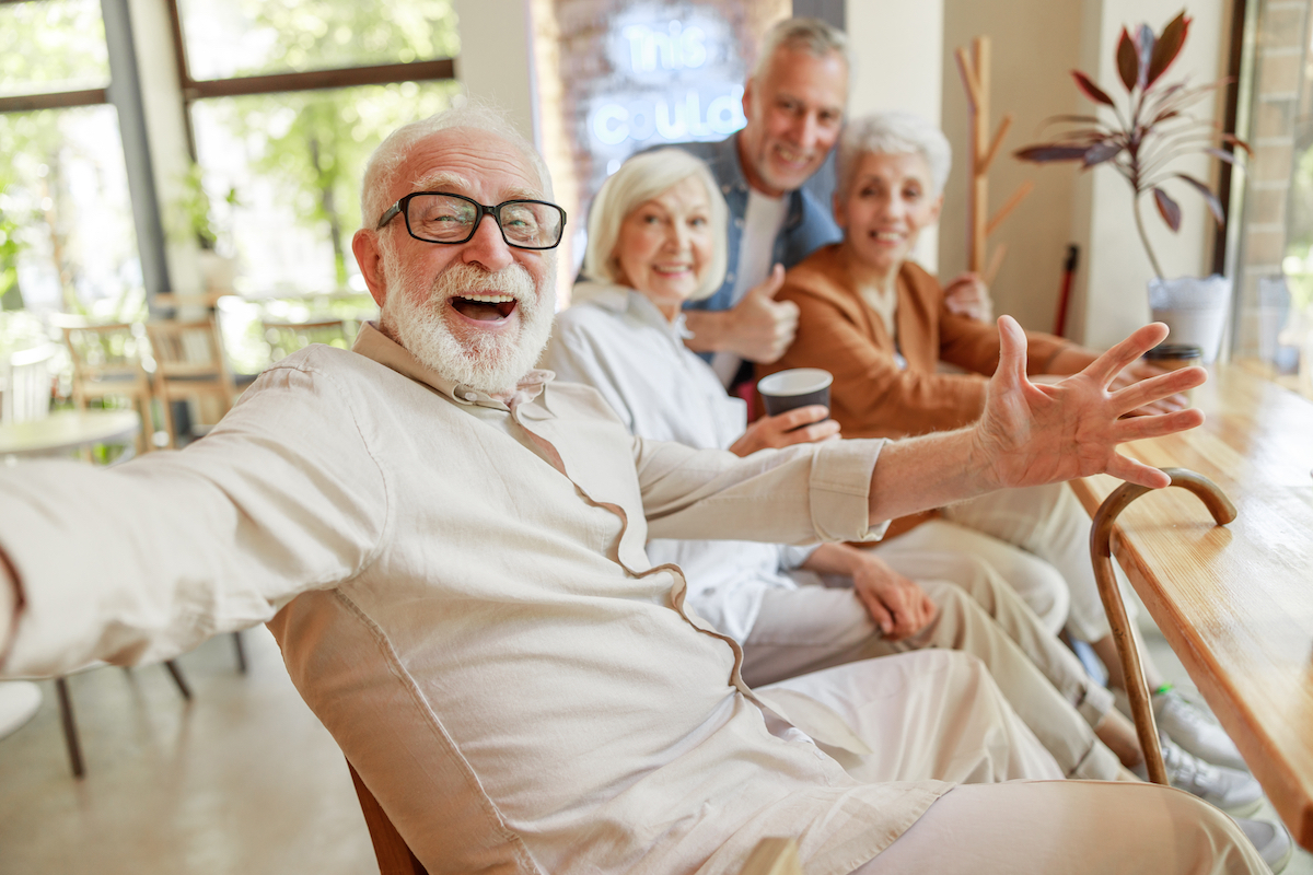 A joyful senior man smiling with his arms spread wide with three friends in the background.