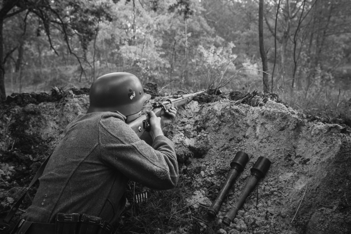 Unidentified Re-enactor Dressed As German Wehrmacht Infantry Soldier In World War II Hidden Sitting With Rifle Weapon In An Ambush In Trench In Autumn Forest. Photo In Black And White Colors.