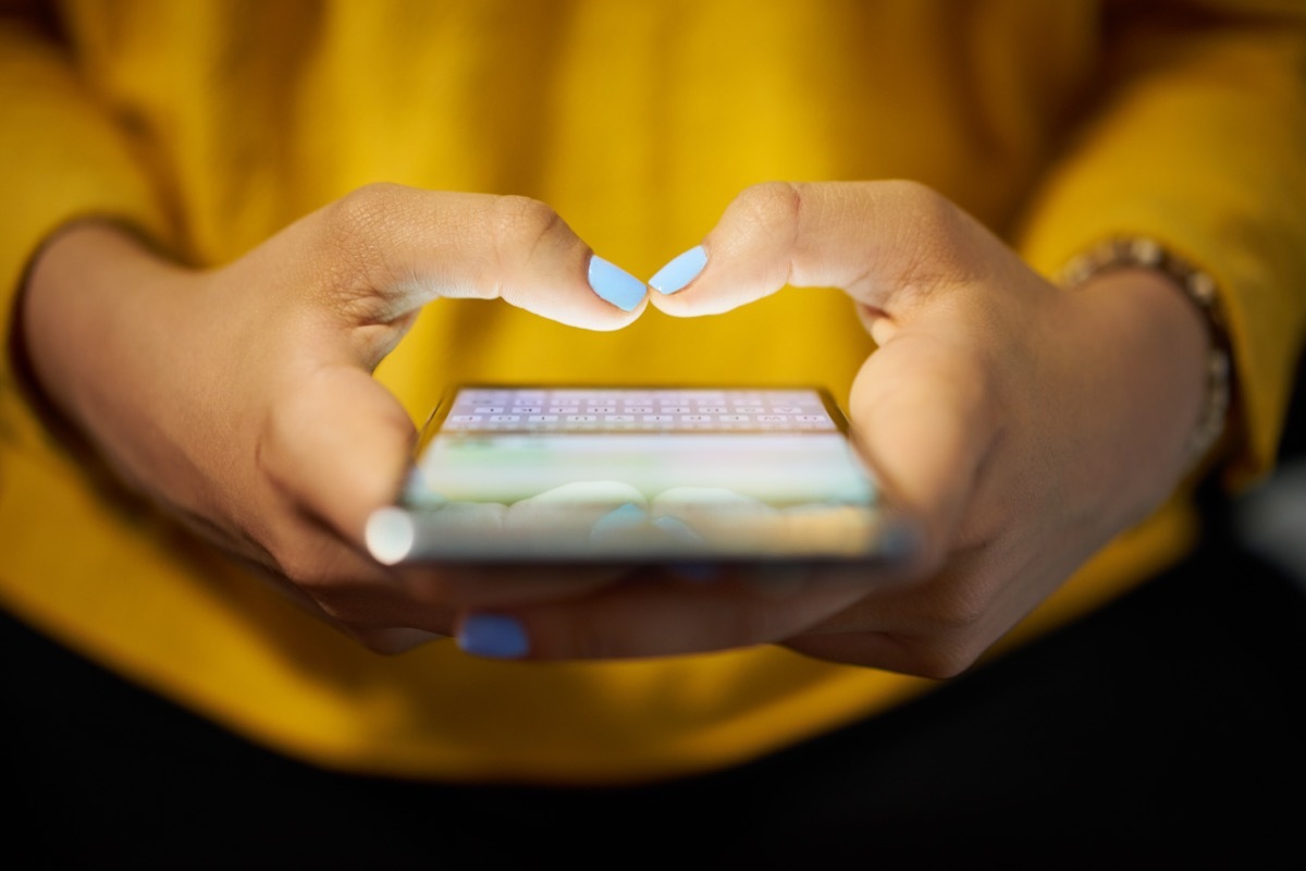 Young woman using cell phone to send text message on social network at night. Closeup of hands with computer laptop in background