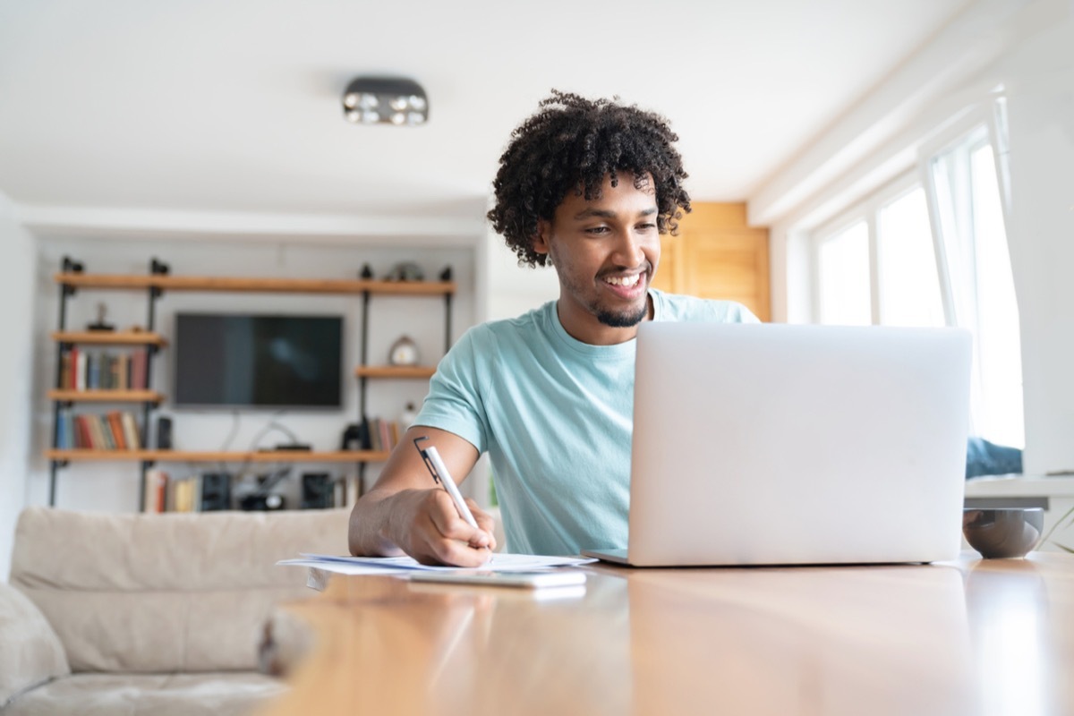 young black man smiling while writing something down and looking at his laptop