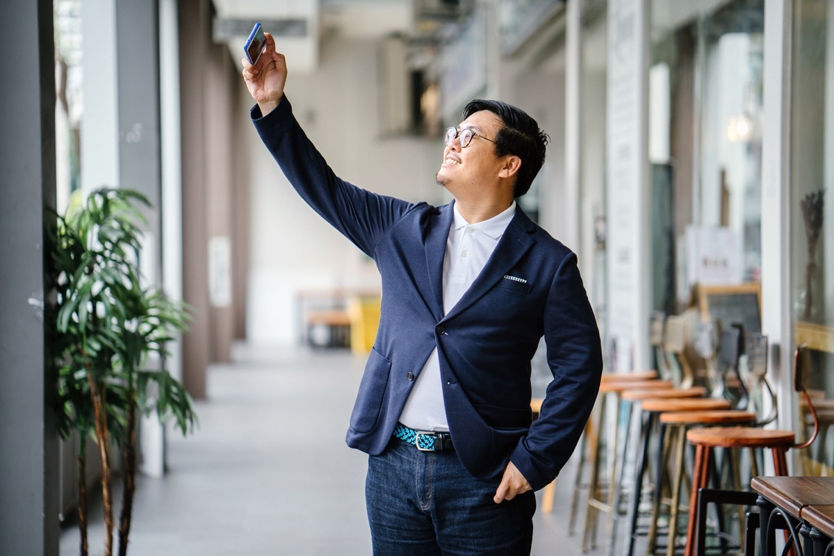 man wearing a blue blazer and dark jeans holding a smartphone and talking a selfie