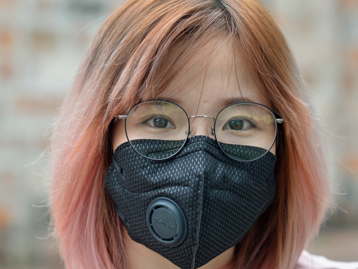 woman with dyed hair and eyeglasses wears a black protective face mask with breathing valve.