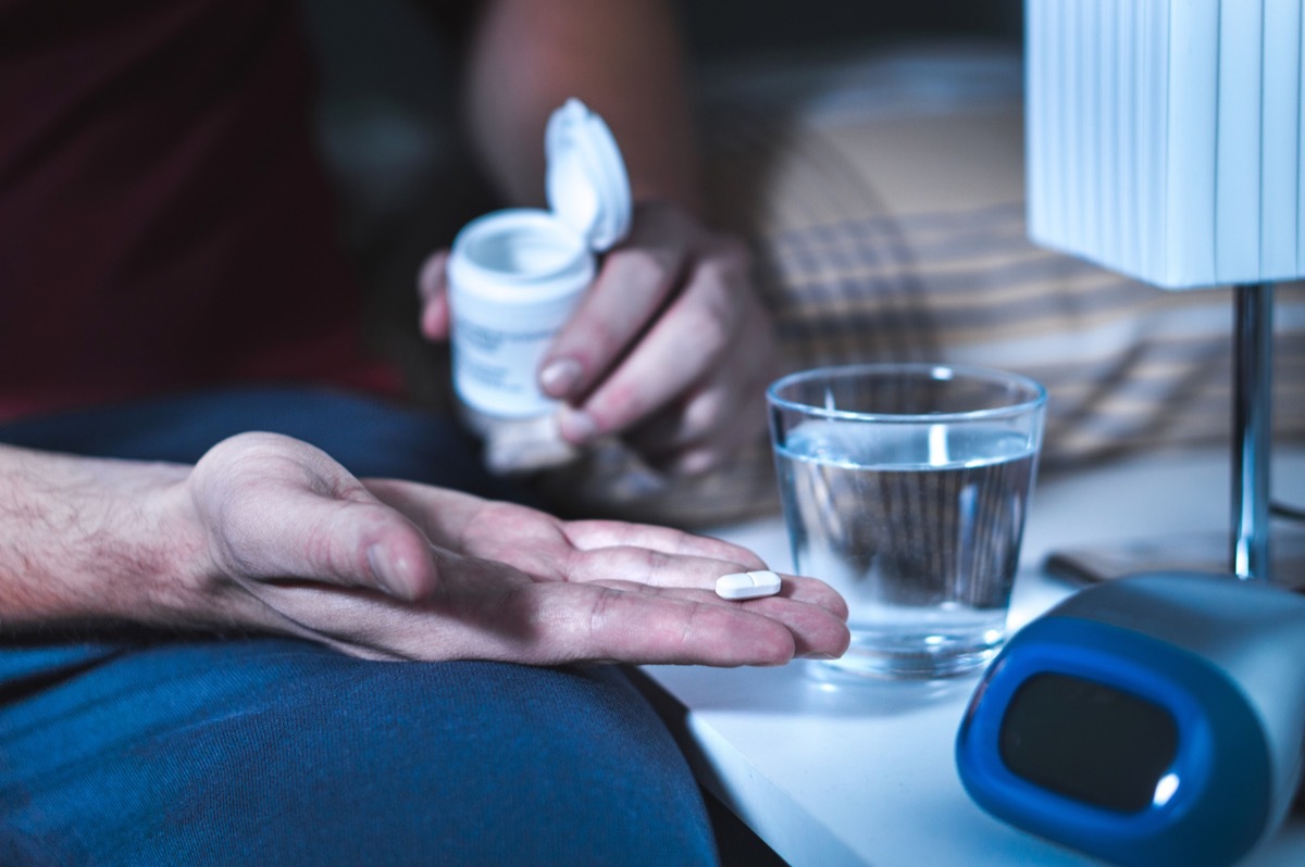 Man sitting on bed about to take sleeping pill or night medicine. Suffering from insomnia. Holding tablet and bottle in hand. Glass of water on nightstand in bedroom.