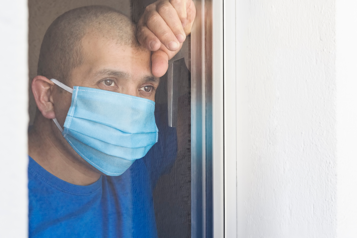 Sick man with face mask looking out the window being quarantined at home.