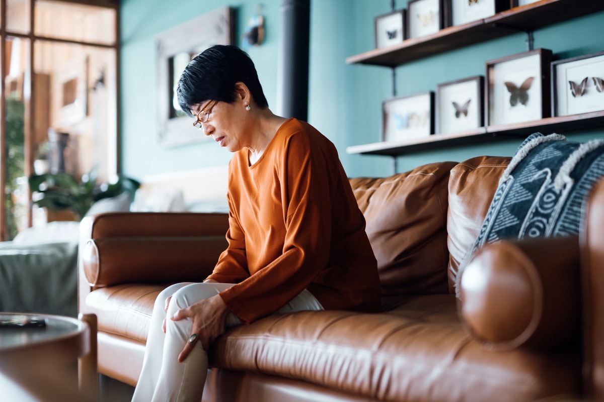 woman feeling unwell, suffering from pain in leg while sitting on sofa in the living room at home