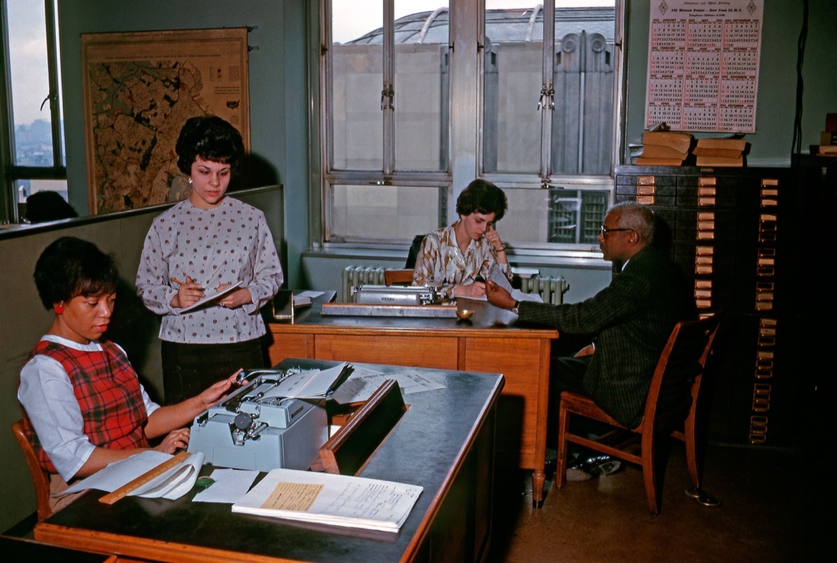 1960s black woman sits at typewriter in an office with three colleagues, shows how different parenting was in the 1950s