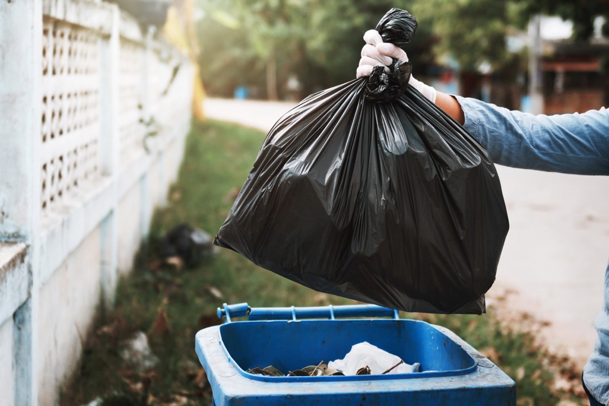 person putting black trash bag into large outdoor trash can