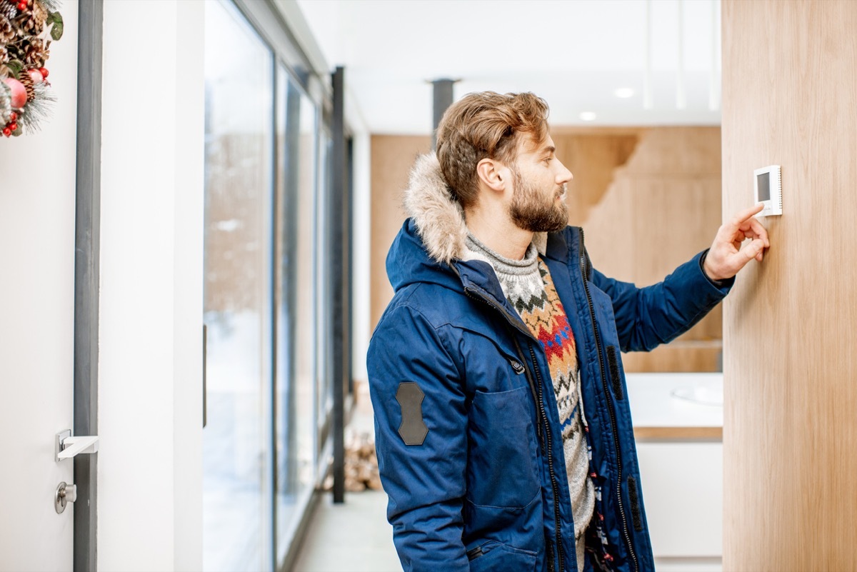 young white man in a winter coat adjusting the thermostat in his home