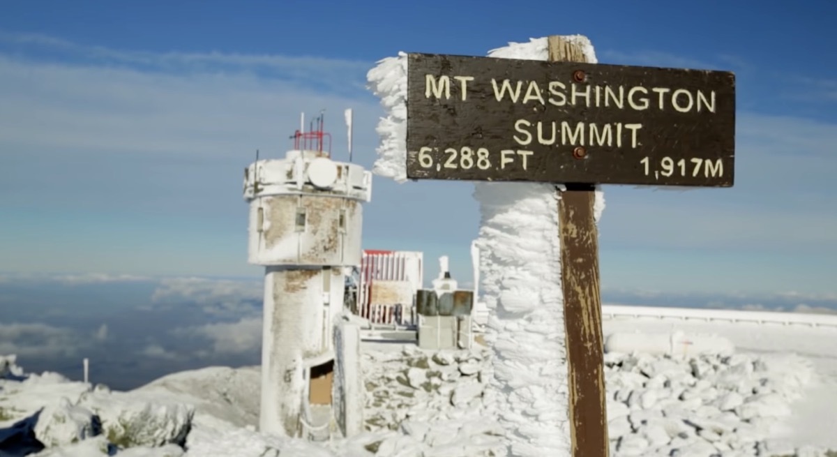 The Mount Washington Observatory summit with a sign covered in snow that reads an elevation of 6,220 feet