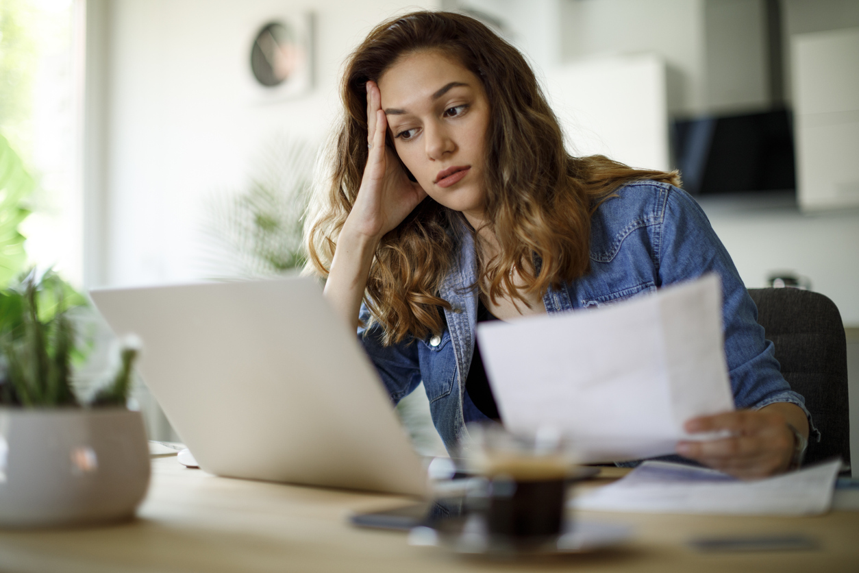 A woman using her laptop while looking over tax information or bills with a concerned look on her face