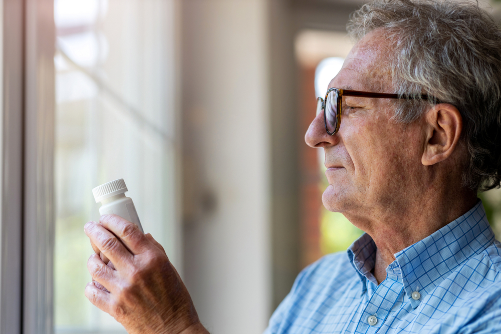 A senior man looking at a supplement bottle while standing near a window