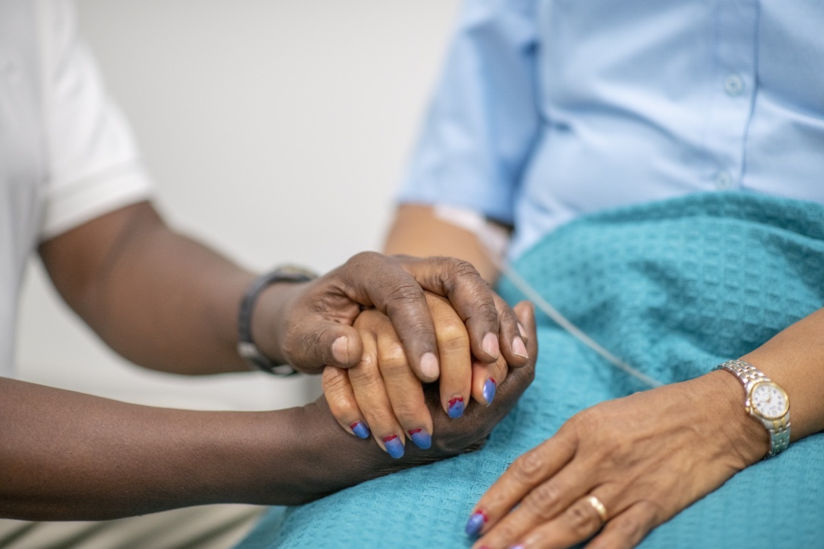 Elderly woman is comforted by a medical professional during the Covid-19. Focus is on their hands. The medical staff is holding the woman's hand.