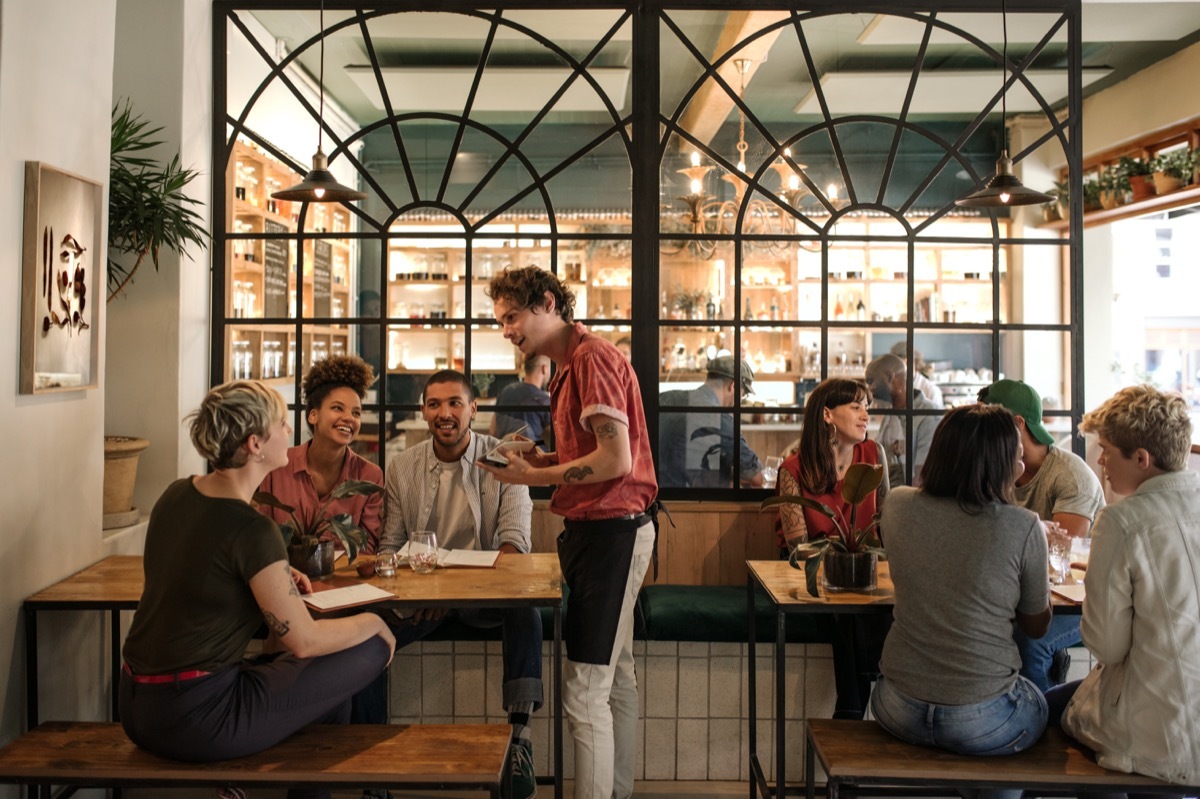 Smiling young waiter taking orders from a diverse group of customers sitting together at a restaurant table