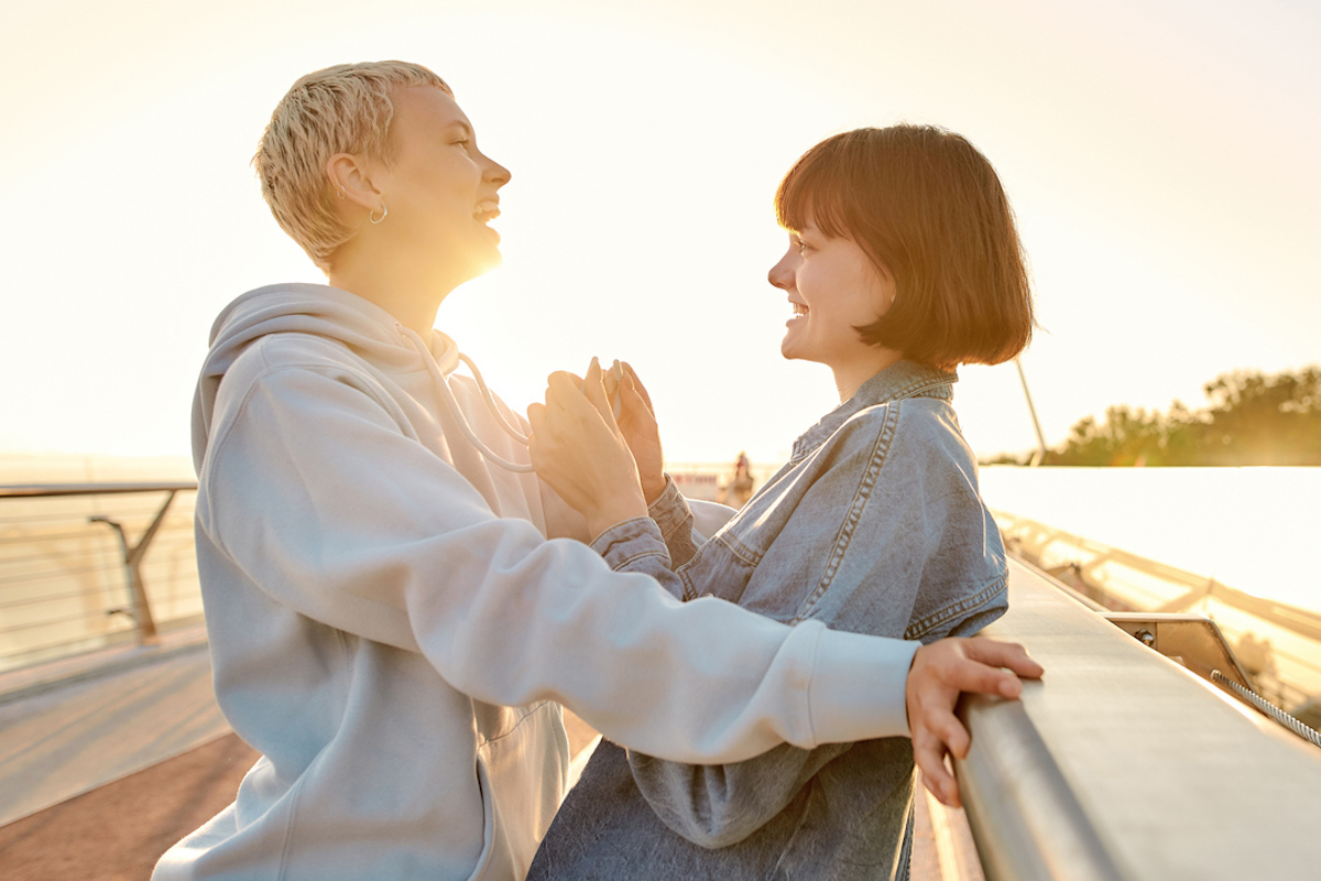 two women looking at each other, laughing together while leaning on the bridge and watching the sunrise