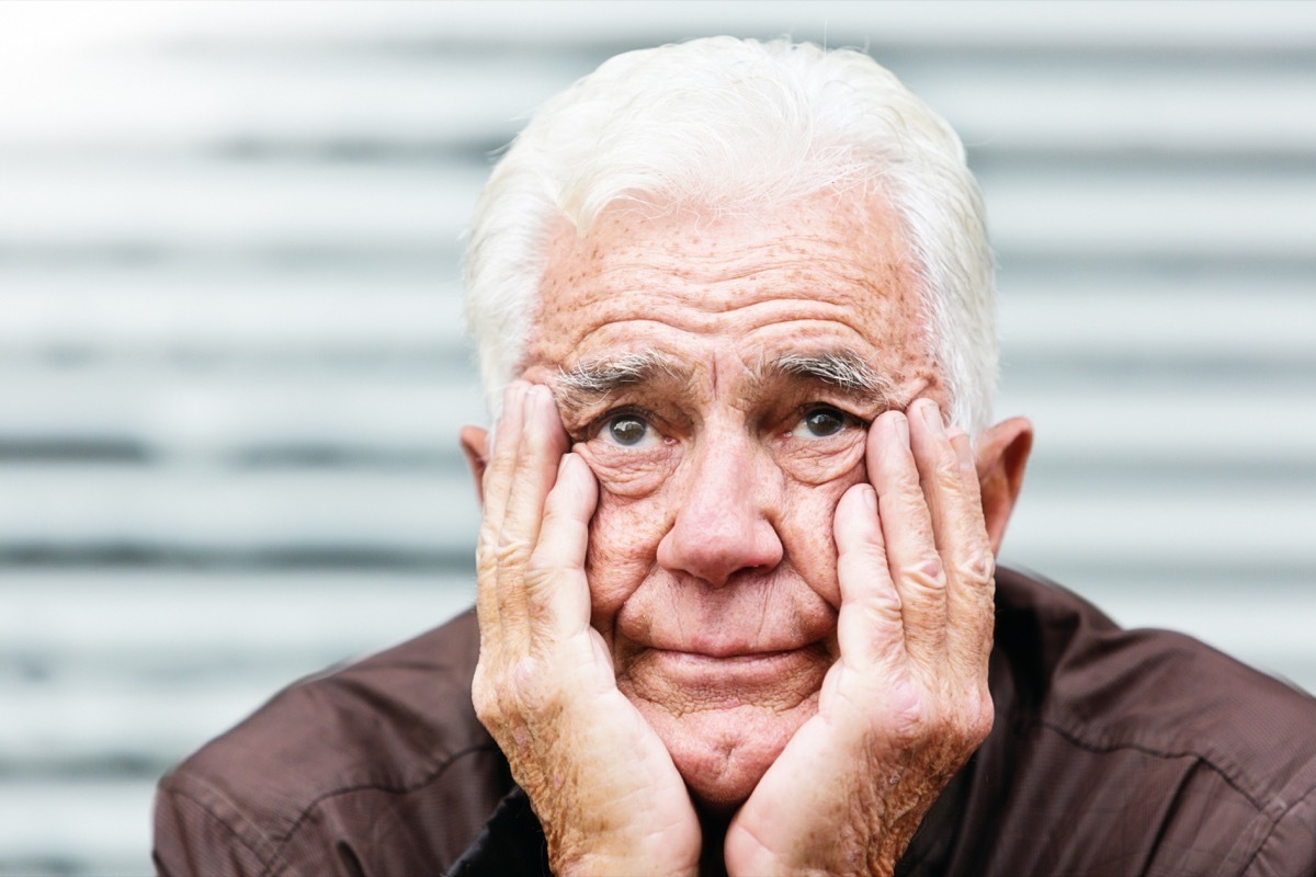 An unhappy old man, his head in his hands looks at camera, frowning.