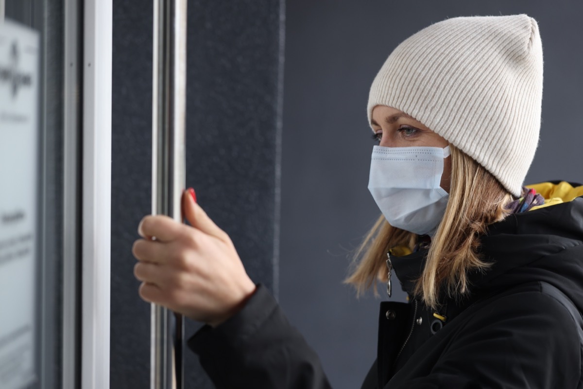 Young woman in protective medical mask and warm clothing opening door to store