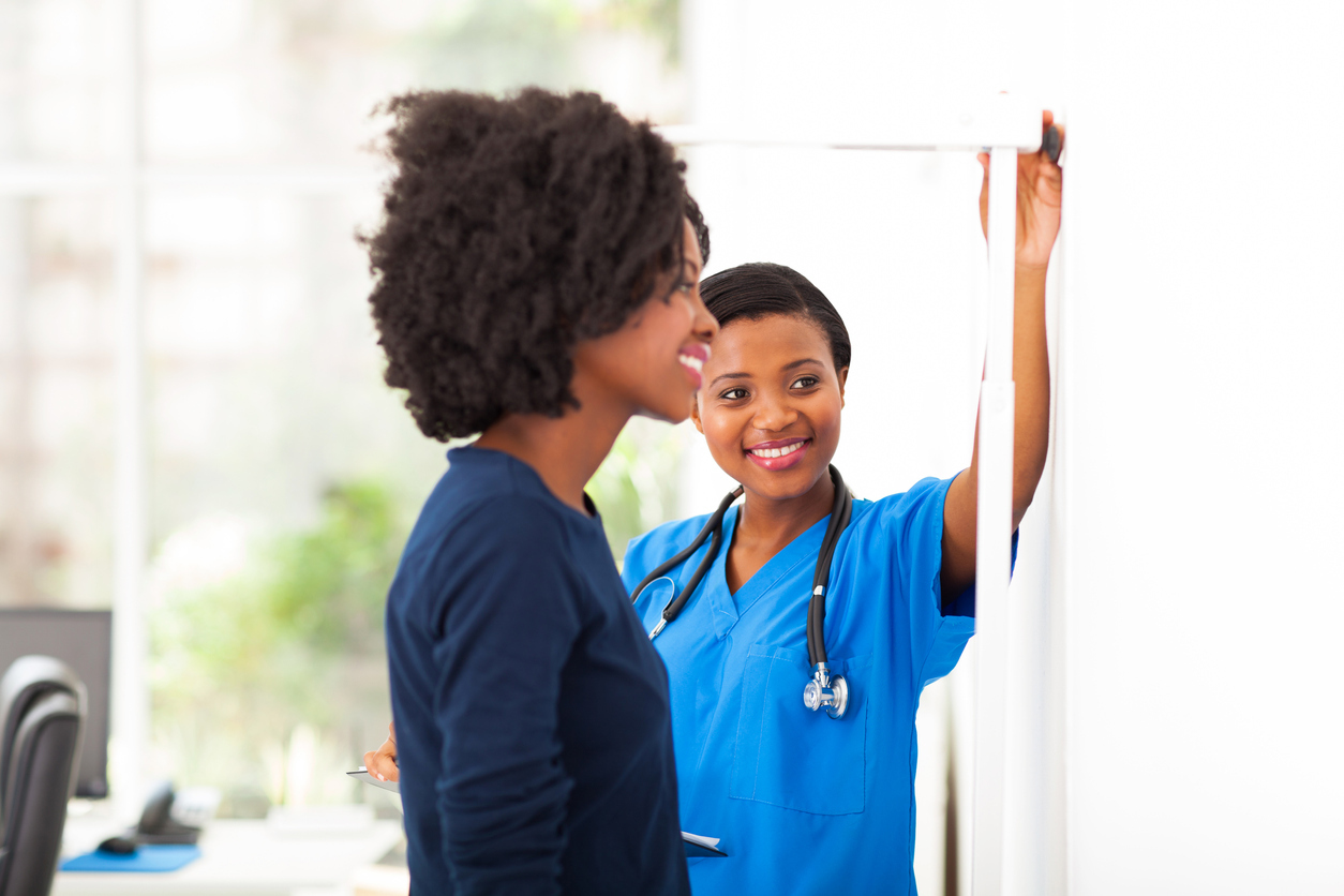 A Black nurse measuring the height of a Black female patient to see how tall she is