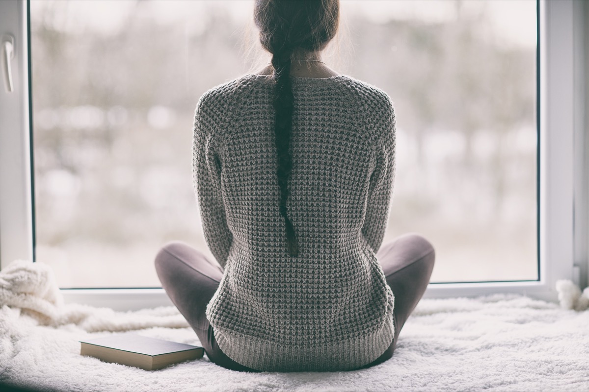 Thoughtful young brunette woman with book looking through the window, blurry winter forrest landscape outside