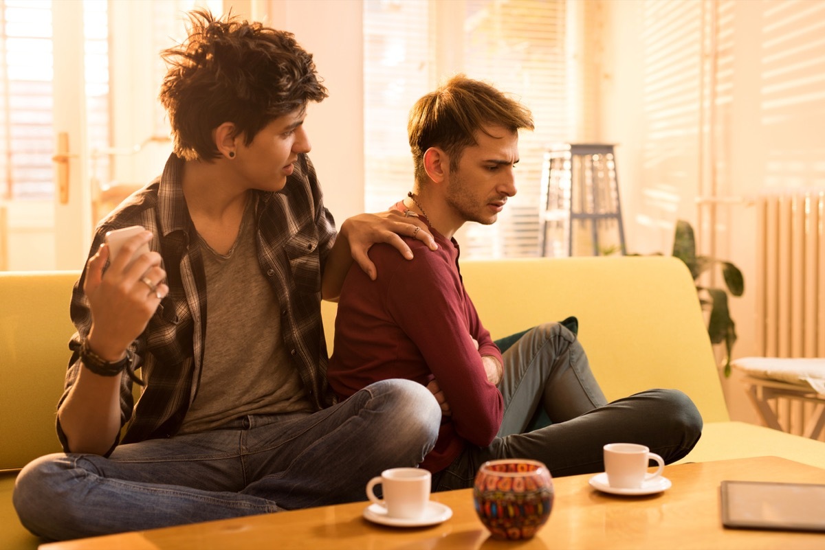 Young gay man puts hand on shoulder of boyfriend or husband who has his back turned while they sit on couch