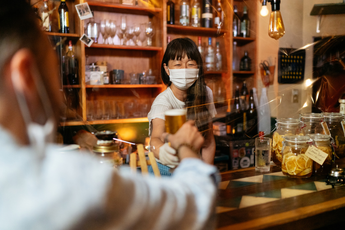 A bar owner is giving beer to customer from behind a protective clear plastic curtain while wearing a mask