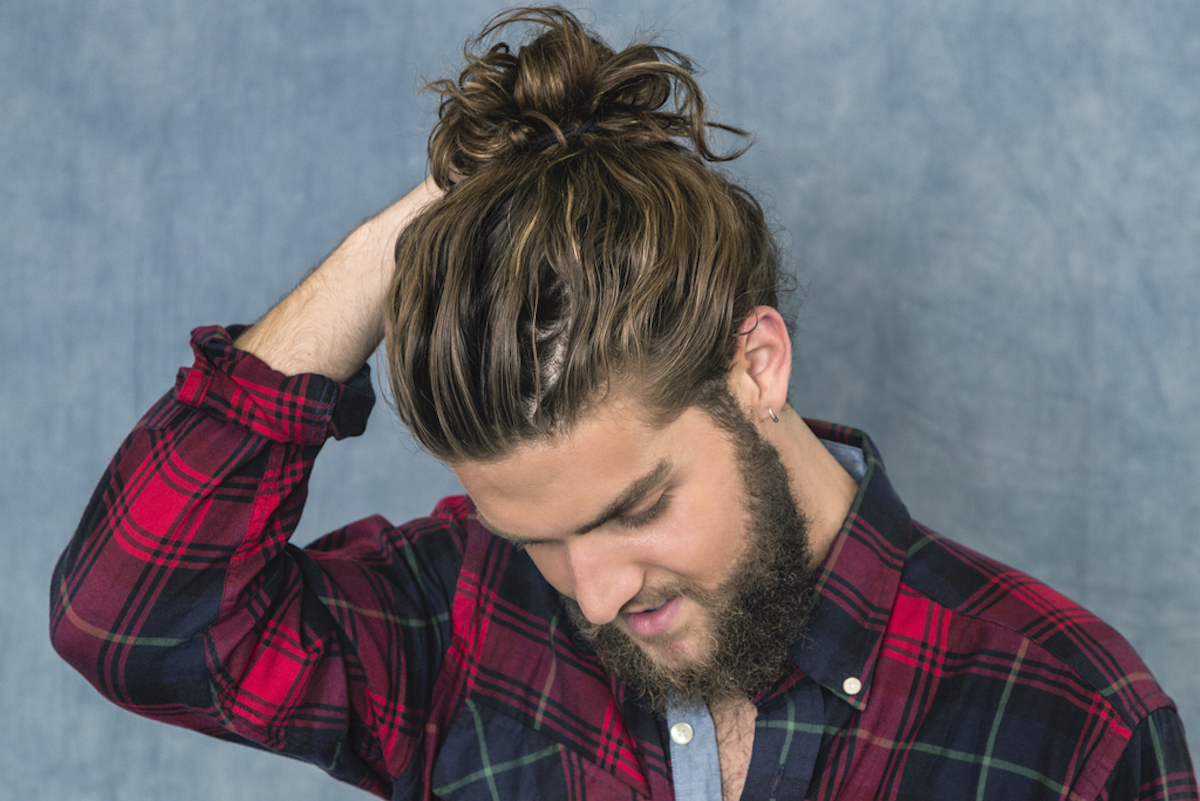 Portrait of a young man with long, tousled brown hair and a full beard wearing a plaid lumberjack shirt and adjusting his bun