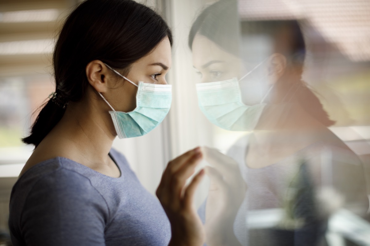 Sad woman with face protective mask looking through the window at home.