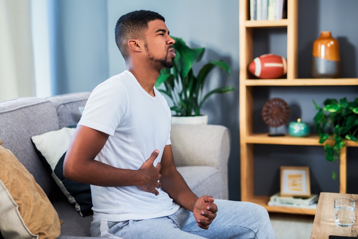 Shot of a young man suffering from stomach pain while lying sitting a sofa at home