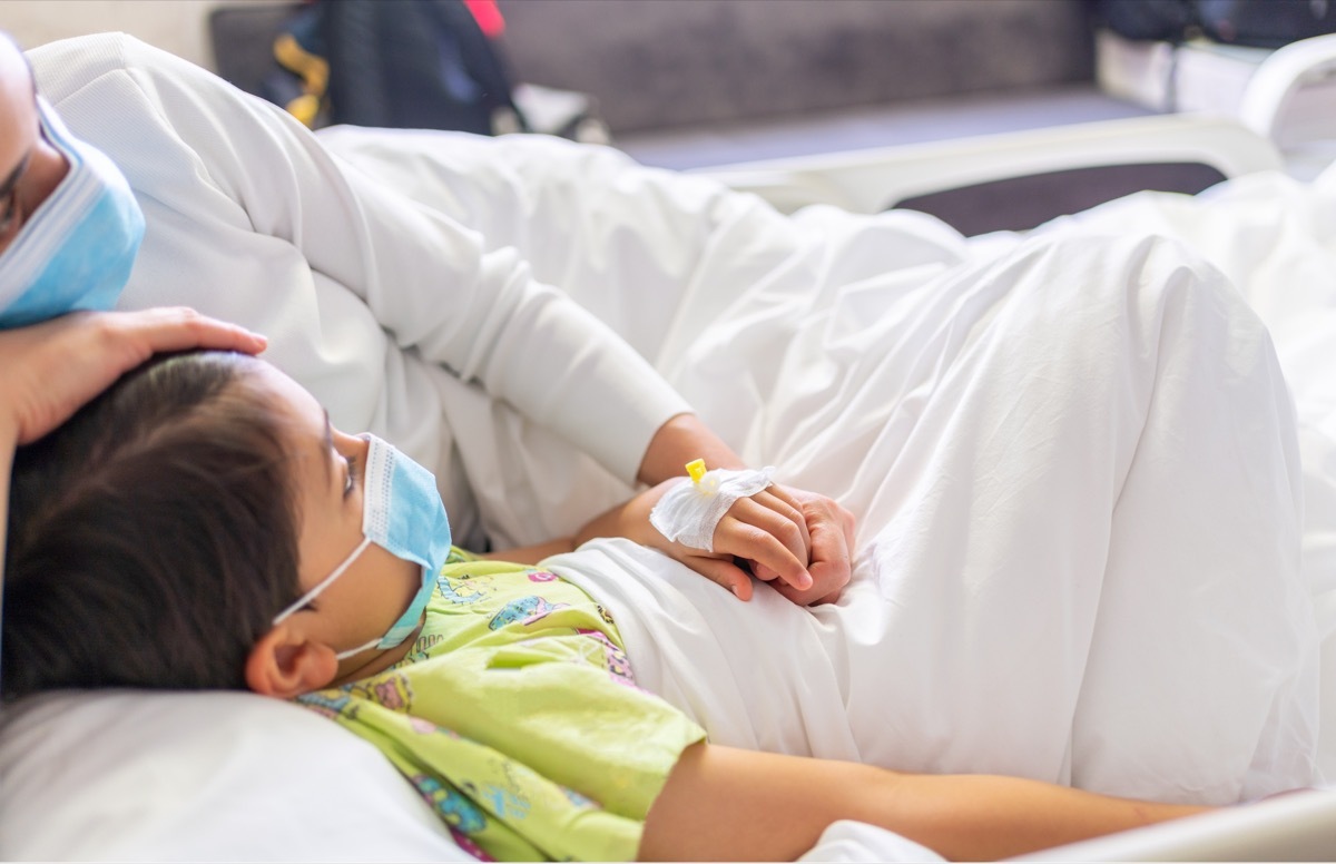 Young mother with protective face mask holding son's hand while lying on the hospital bed with him. Emotional Family Moment. Child vascular access