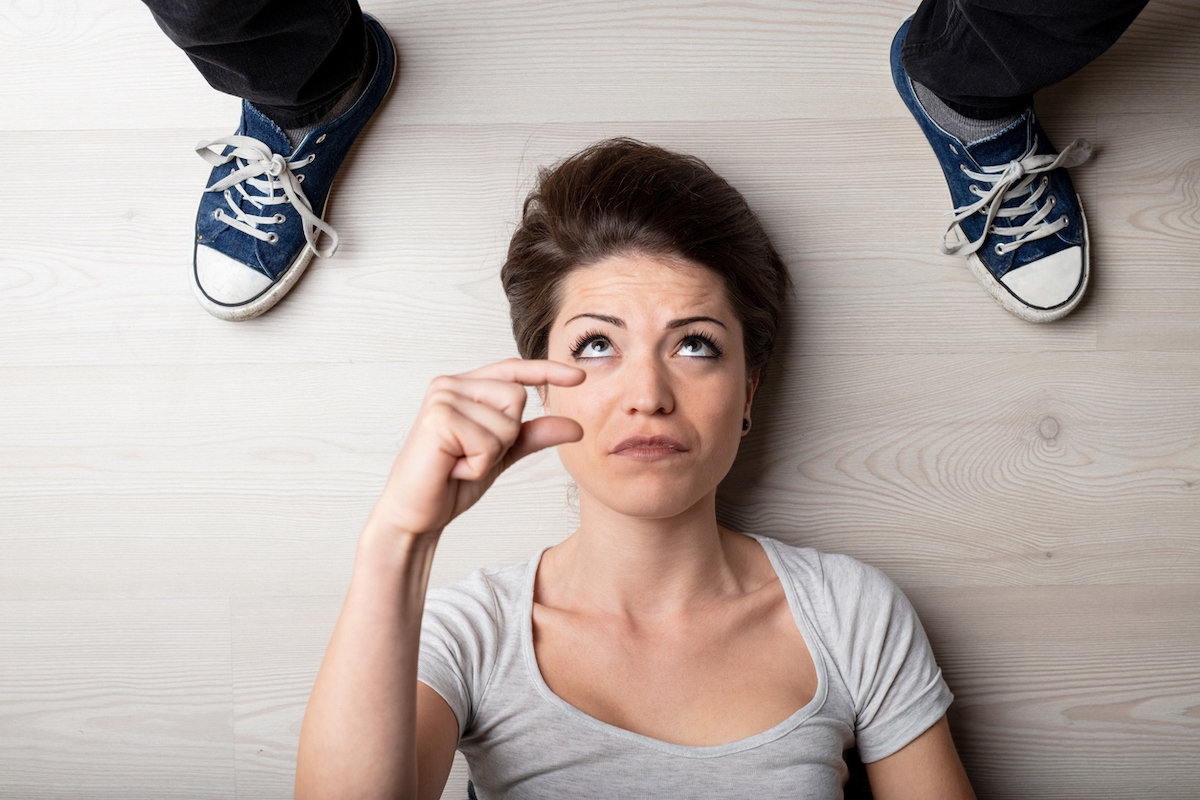 Woman making a small or short gesture with her fingers as she lies on the floor between the feet of a person wearing jeans and sneakers