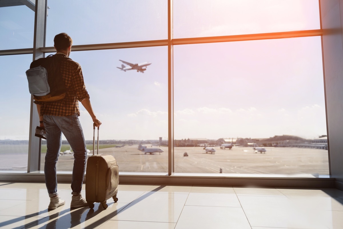 male tourist is standing in airport and looking at aircraft flight through window. He is holding tickets and suitcase. Sunset