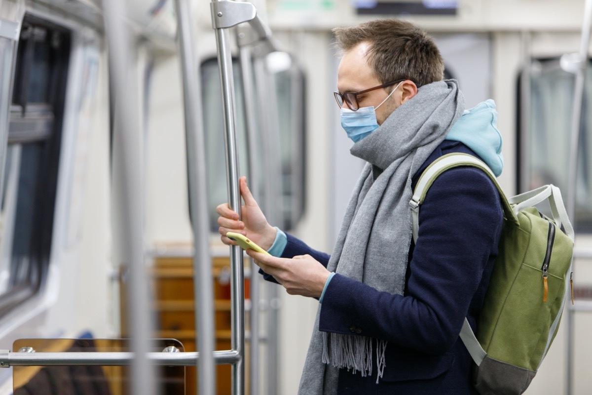 man in glasses feeling sick, wearing protective mask against transmissible infectious diseases and as protection against the flu in public transport/subway, using and looking at smartphone