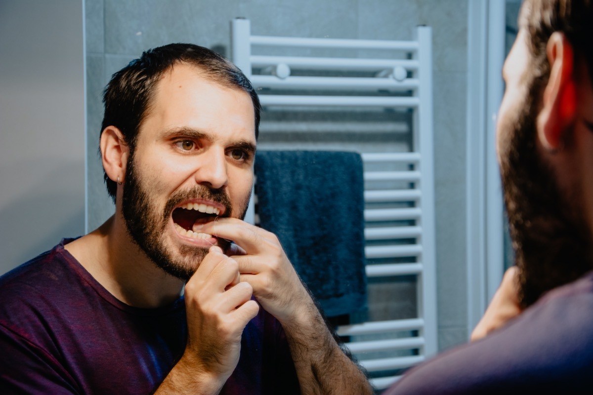 cleaning his teeth with dental floss and smiling while standing in front of the mirror