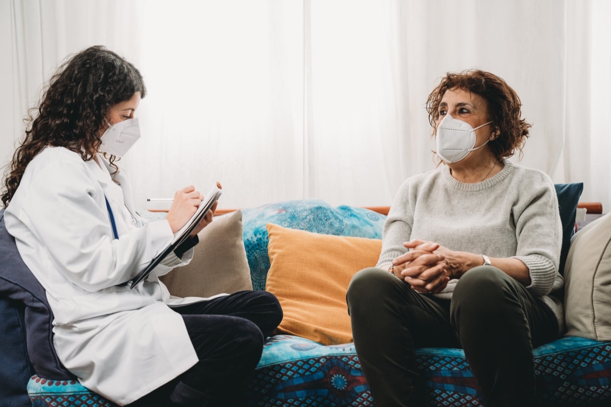 Female doctor is visiting a patient at home, holding a notebook. She's asking questions to a senior woman.