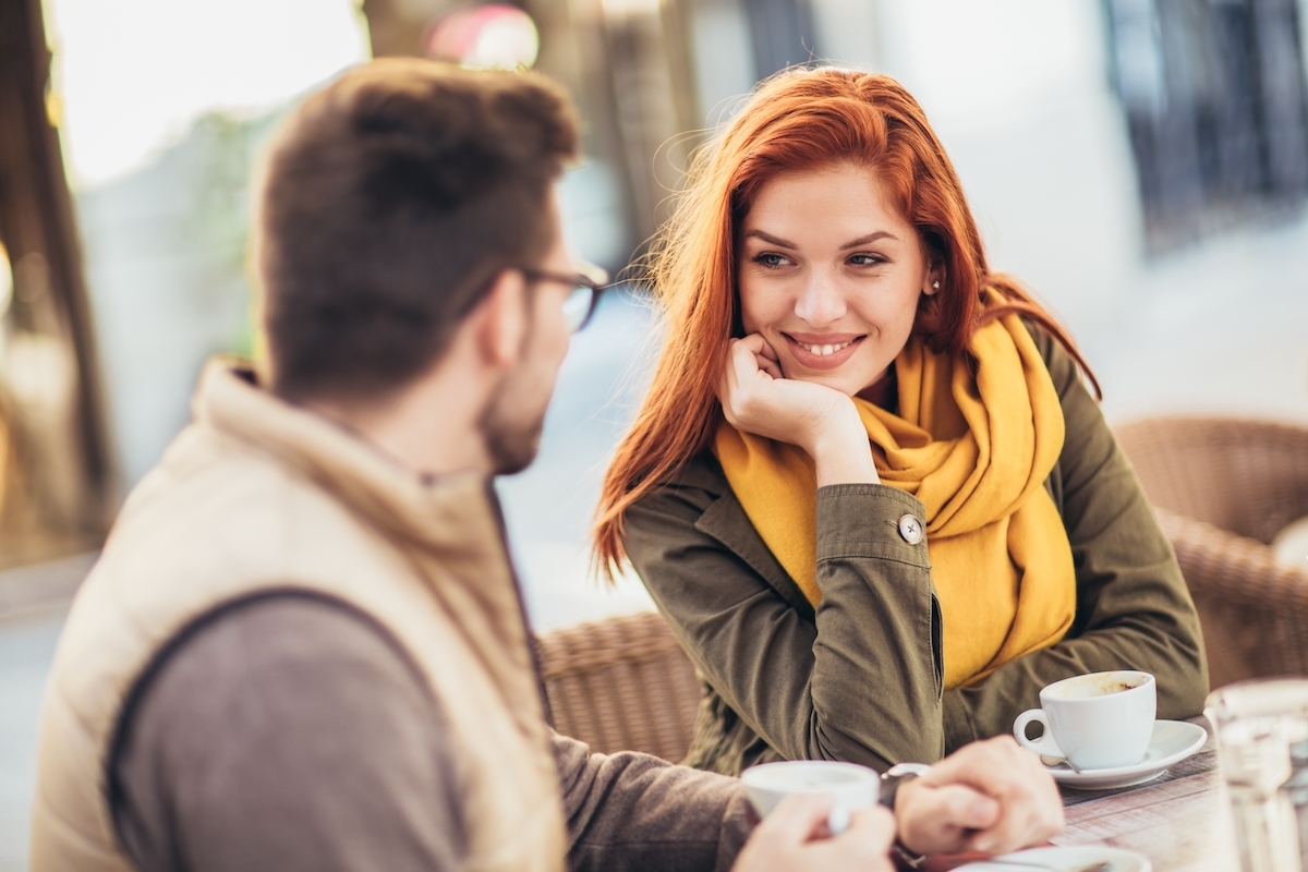 A couple sitting at an outdoor cafe; the woman is smiling and staring at her partner.