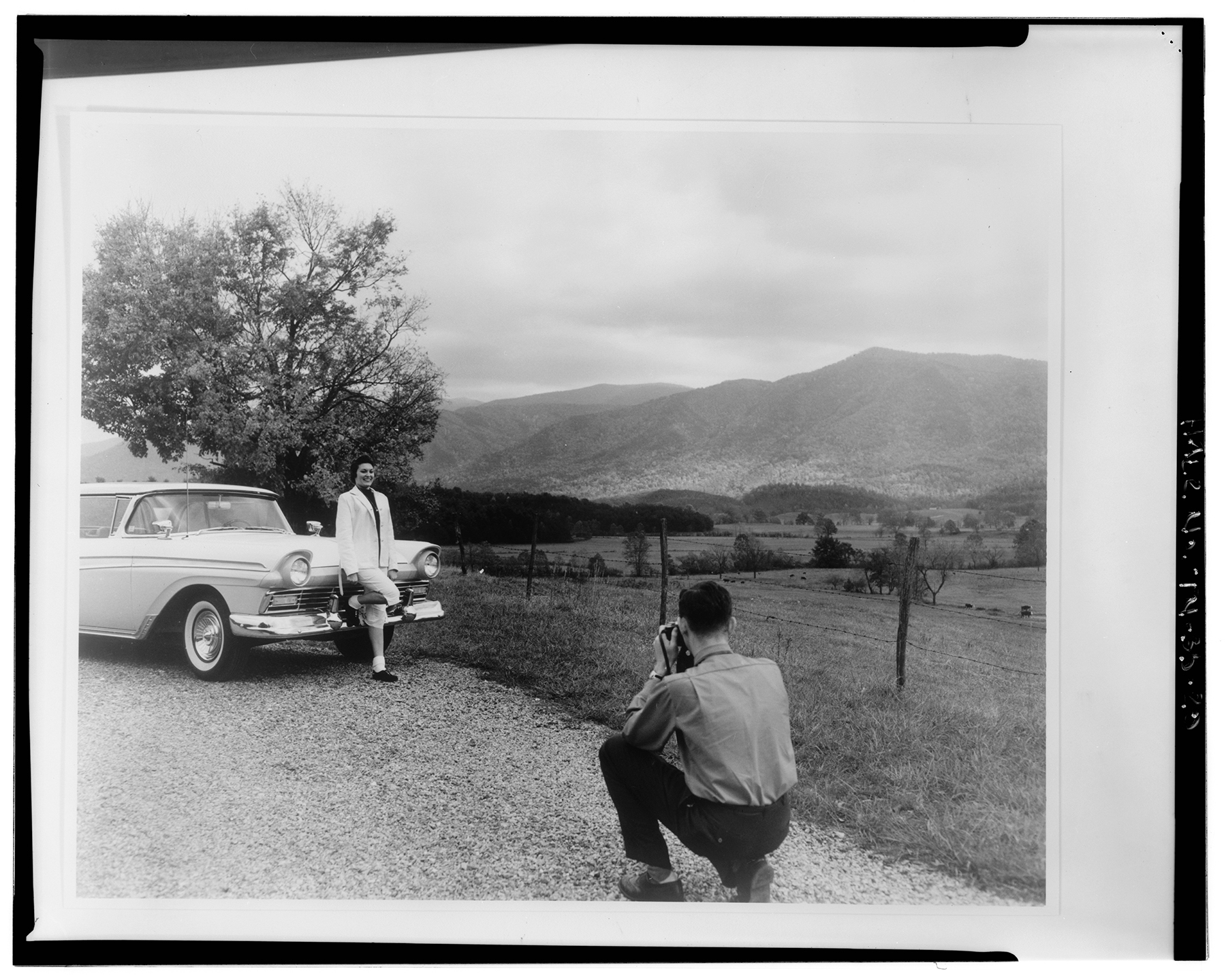 a man takes a photo of a girl leaning against a car