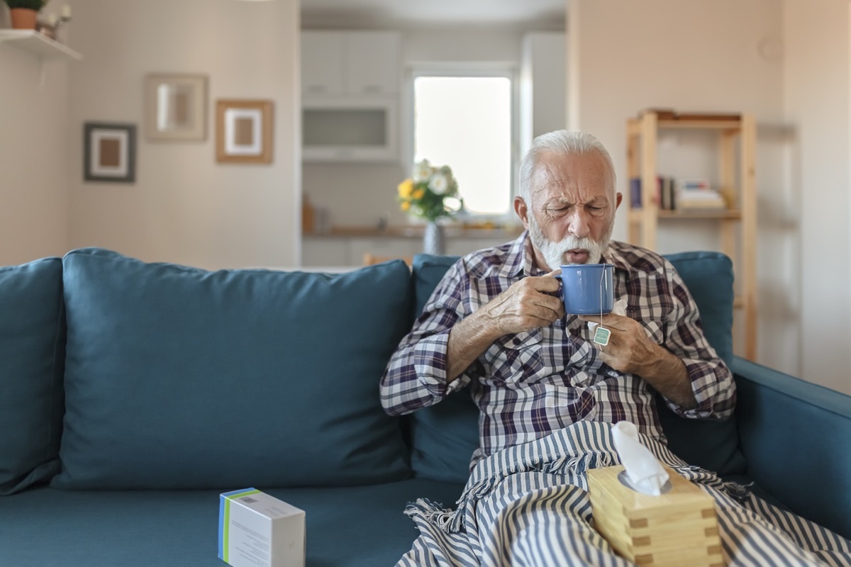 Senior man suffering from flu drinking tea while sitting wrapped in a blanket on the sofa at home. Sick older man with headache sitting under the blanket in the living room. Man with a cold lying on the sofa holding a mug of hot tea.