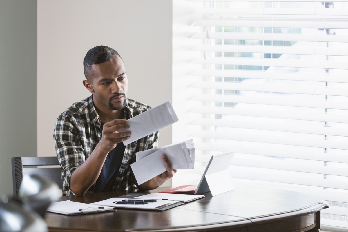 man sitting at a desk by a window at home, paying bills. He is looking through a stack of envelopes and has his laptop computer in front of him.