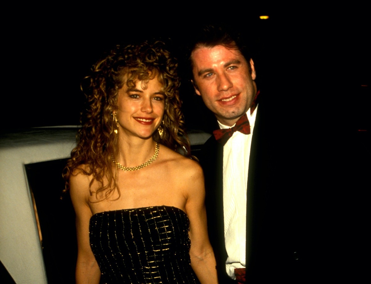 John Travolta wears a black suit and white shirt and Kelly Preston wears a black dress outside of a restaurant in 1991