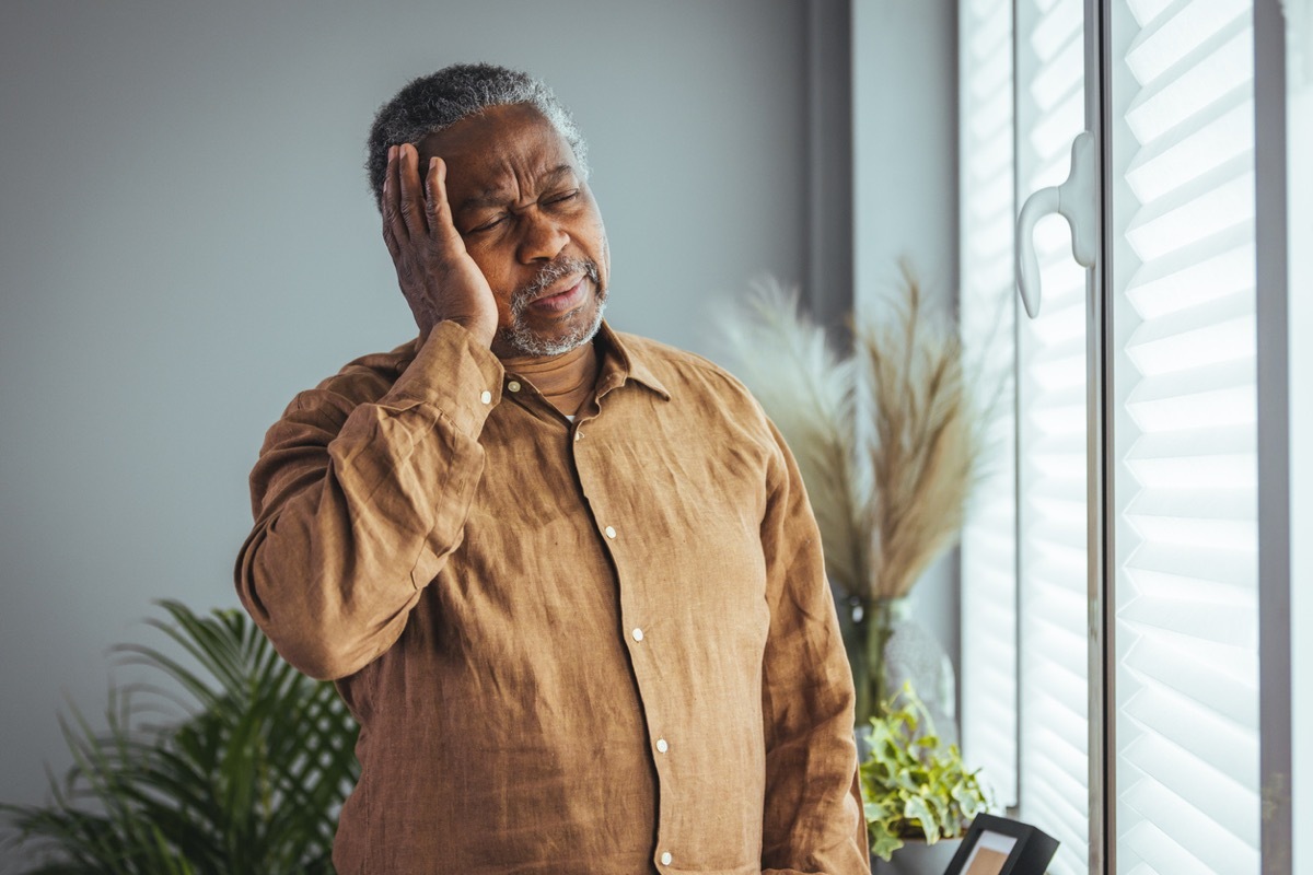 An older, gray-haired man touches his head looking distressed while standing near a window