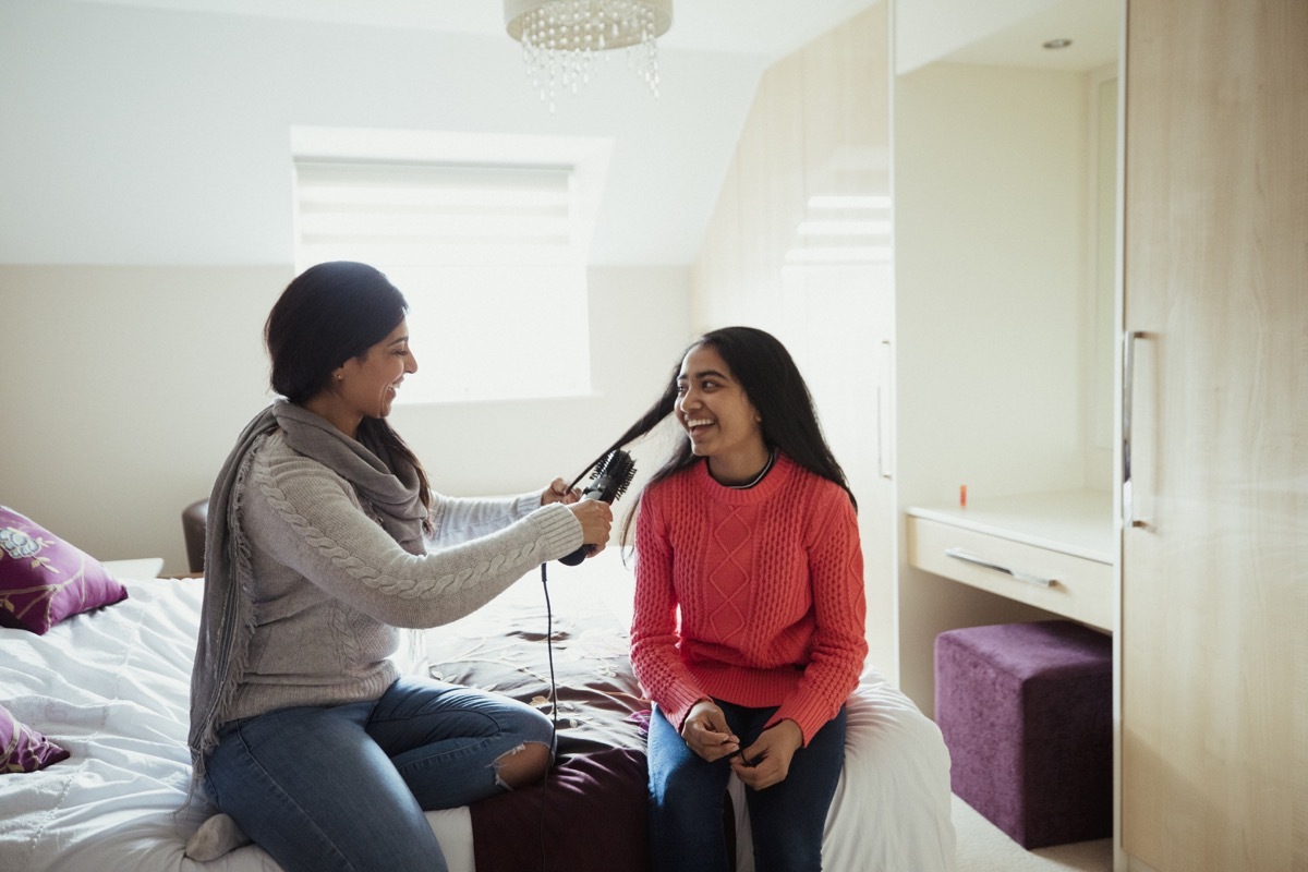 A mother and her daughter are sitting on her bed whilst she curls her daughter's hair. They both look happy to be bonding and sharing this moment together.