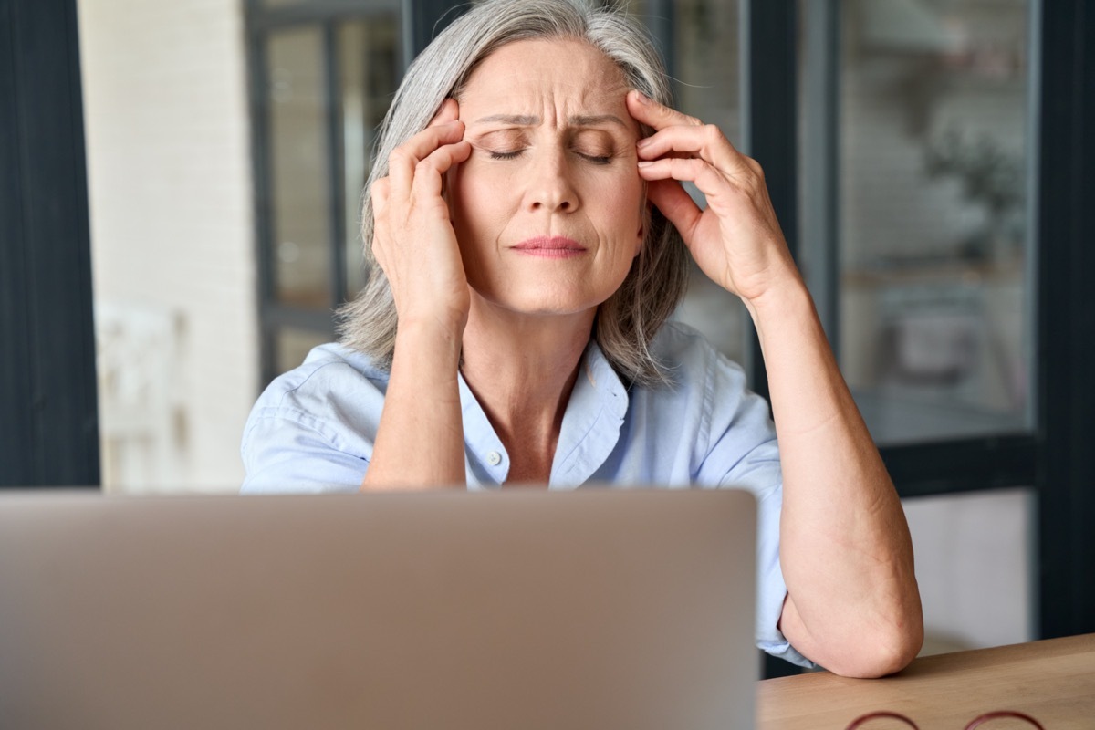 Stressed middle 60s aged worker woman massaging head suffering of headache in home office.